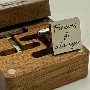 A square silver cufflink with the words "Forever & always" engraved on it, resting on a wooden box. The box is open, revealing a compartment for cufflinks. The wood has a natural grain.