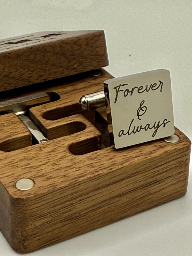 A square silver cufflink with the words "Forever & always" engraved on it, resting on a wooden box. The box is open, revealing a compartment for cufflinks. The wood has a natural grain.