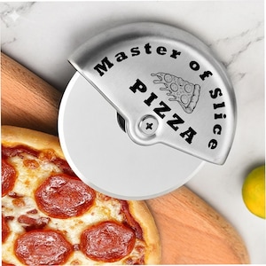 Close-up of a stainless steel pizza cutter engraved with “Master of Slice Pizza” and a pizza slice illustration, positioned over a wooden board beside a pepperoni pizza on a light marble surface.