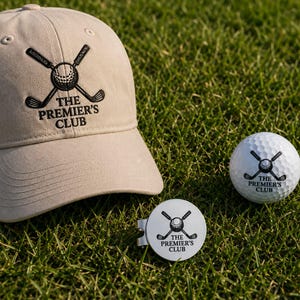 Engraved golf hat and custom ball marker set with clip. Personalized groomsmen golf gifts featuring "The Premier's Club" logo with crossed golf clubs on grass.