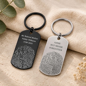 Two personalized fingerprint memorial keychains displayed on a neutral fabric background with eucalyptus leaves and dried flowers. The black keychain is engraved with “My Beloved Mother Ashley Williams 1938–2025,” while the silver keychain features the message “Gone But Never Forgotten.” Both designs include detailed fingerprint engravings, showcasing custom remembrance keepsakes for honoring loved ones.
