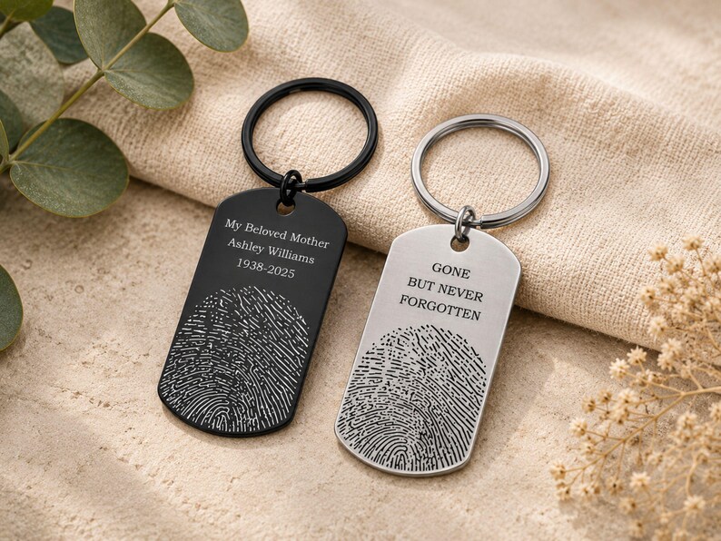 Two personalized fingerprint memorial keychains displayed on a neutral fabric background with eucalyptus leaves and dried flowers. The black keychain is engraved with “My Beloved Mother Ashley Williams 1938–2025,” while the silver keychain features the message “Gone But Never Forgotten.” Both designs include detailed fingerprint engravings, showcasing custom remembrance keepsakes for honoring loved ones.