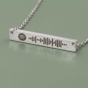 Silver-colored necklace featuring a rectangular pendant with a Spotify code and logo. The pendant is connected to a delicate chain. The necklace is displayed on a white surface.