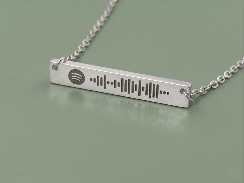 Silver-colored necklace featuring a rectangular pendant with a Spotify code and logo. The pendant is connected to a delicate chain. The necklace is displayed on a white surface.