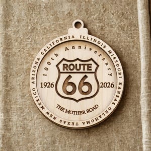 Op de afbeelding: Een ronde houten Route 66 jubileumornament. Het ontwerp toont het Route 66-schild met de nummers 66, de jaren 1926 en 2026, en de tekst "100th Anniversary". De staten staan rond de rand.