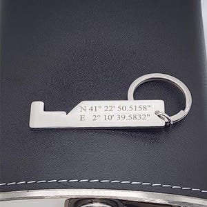 A black leather flask with a silver-colored key chain. The key chain has engraved coordinates: N 41° 22' 50.5158" E 2° 10' 39.5832". The flask has a silver cap and trim with white stitching.