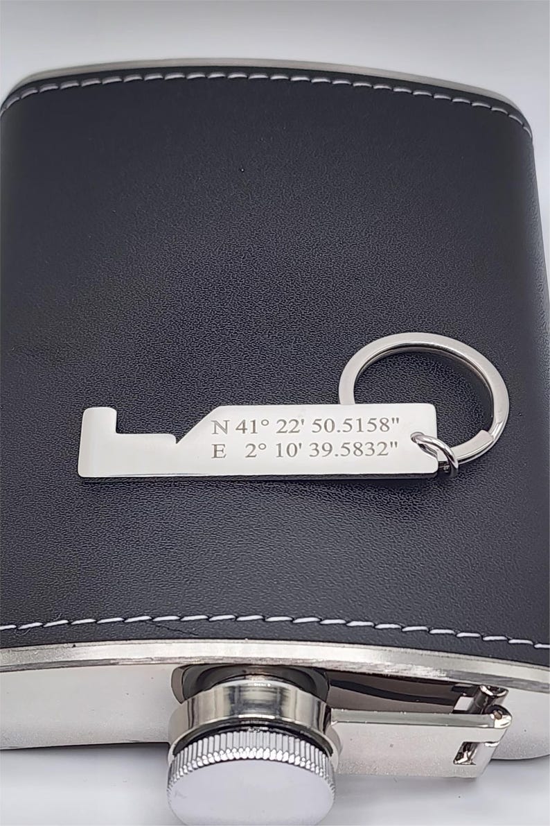 A black leather flask with a silver-colored key chain. The key chain has engraved coordinates: N 41° 22' 50.5158" E 2° 10' 39.5832". The flask has a silver cap and trim with white stitching.
