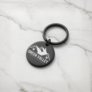 May include: A black round keychain with a black key ring. The keychain has the words "Green Valley Premium Properties" and a house graphic in white. The keychain is on a white and gray marble surface.