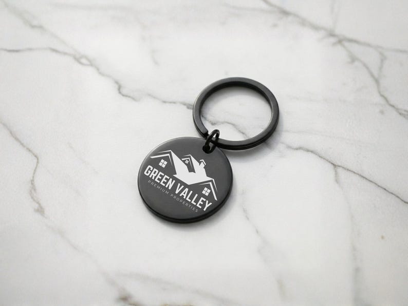 May include: A black round keychain with a black key ring. The keychain has the words "Green Valley Premium Properties" and a house graphic in white. The keychain is on a white and gray marble surface.