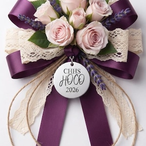 K&ouml;nnte beinhalten: Ein Blumen-Corsage mit hellrosa Rosen, gr&uuml;nen Bl&auml;ttern und violetten Lavendelzweigen. Es ist mit dunkelviolettem Satinband und cremefarbener Spitzenborte verziert. Ein runder silberner Anh&auml;nger in der Mitte tr&auml;gt die Aufschrift CHHS HOCO 2026.