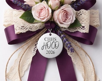 May include: A floral corsage featuring light pink roses, green leaves, and purple lavender sprigs. It is accented with deep purple satin ribbon and cream-colored lace trim. A circular silver charm hangs from the center, inscribed with CHHS HOCO 2026.