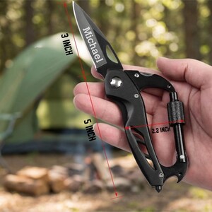 A hand is holding a black carabiner multi-tool, which is made of metal and plastic. The multi-tool has several features, including a bottle opener, a wrench, a Phillips screwdriver, and a flathead screwdriver. The tool is held against a background of a forest trail. The forest is dense with trees and green foliage. The trail is unpaved and looks like a hiking path. The multi-tool is the main focus of the image, and the background is slightly blurred. The lighting is natural and diffuse.