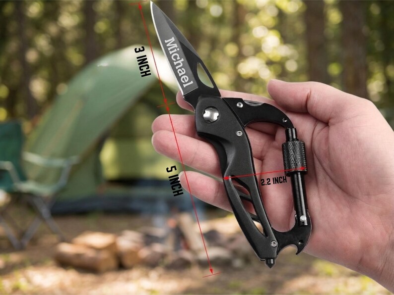 A hand is holding a black carabiner multi-tool, which is made of metal and plastic. The multi-tool has several features, including a bottle opener, a wrench, a Phillips screwdriver, and a flathead screwdriver. The tool is held against a background of a forest trail. The forest is dense with trees and green foliage. The trail is unpaved and looks like a hiking path. The multi-tool is the main focus of the image, and the background is slightly blurred. The lighting is natural and diffuse.