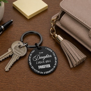 Black round engraved keychain attached to a set of keys on a wooden desk, featuring the message “To my Daughter I love you forever” with a circular motivational quote around the edge. Nearby are a beige purse with a tassel, a black pen, sticky notes, and a small potted plant.
