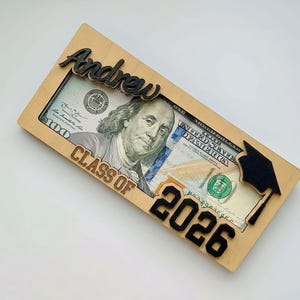 Flat lay of a personalized laser-engraved birch wood graduation money holder reading "Andrew – Class of 2026," with a $100 bill displayed inside the open slot, featuring a raised black script name across the top, bold black laser-cut "2026" numbers at the bottom, and a laser-cut graduation cap with tassel on the right side, photographed at a slight angle against a clean white background.