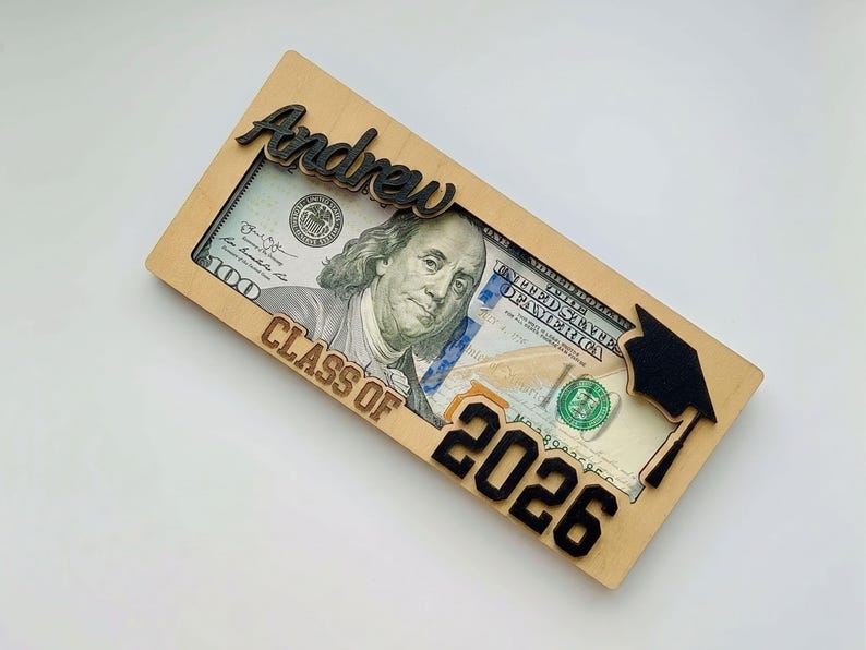 Flat lay of a personalized laser-engraved birch wood graduation money holder reading "Andrew – Class of 2026," with a $100 bill displayed inside the open slot, featuring a raised black script name across the top, bold black laser-cut "2026" numbers at the bottom, and a laser-cut graduation cap with tassel on the right side, photographed at a slight angle against a clean white background.
