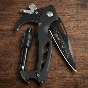 A black multi-tool carabiner knife is centered on a dark, textured wood surface. The knife blade is partially extended, featuring a customizable laser engraving that reads "Your Text Here" in a cursive script. At the top of the handle, a Phillips-head and flathead screwdriver are deployed. The tool also includes a integrated carabiner with a textured locking screw, a bottle opener at the base, and a glass breaker tip. The image is shot from a top-down.