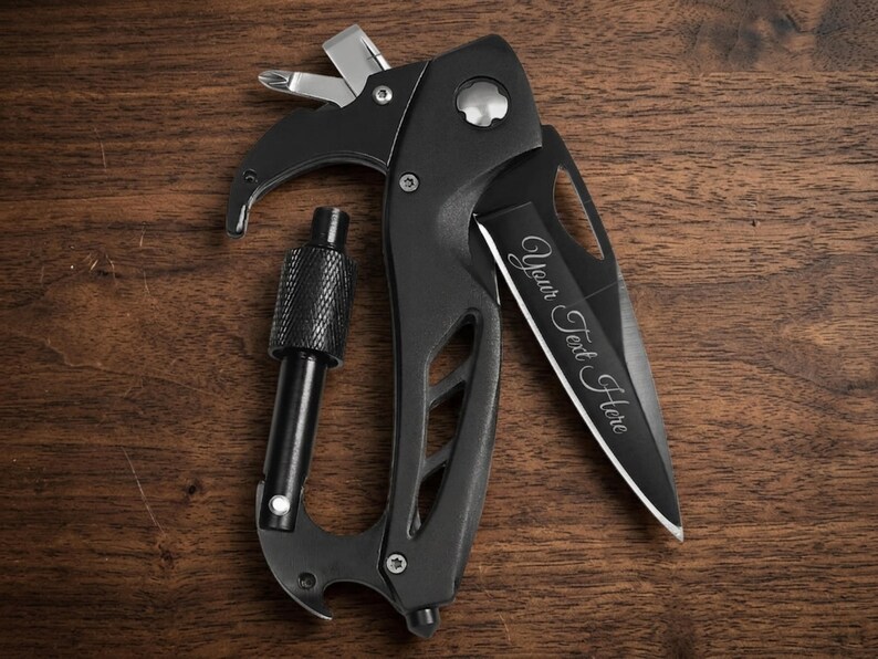 A black multi-tool carabiner knife is centered on a dark, textured wood surface. The knife blade is partially extended, featuring a customizable laser engraving that reads "Your Text Here" in a cursive script. At the top of the handle, a Phillips-head and flathead screwdriver are deployed. The tool also includes a integrated carabiner with a textured locking screw, a bottle opener at the base, and a glass breaker tip. The image is shot from a top-down.