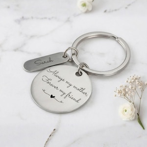 Personalized silver keychain for mom with engraved message “Always my mother, forever my friend” and custom name tag, displayed on a marble background with delicate white flowers.