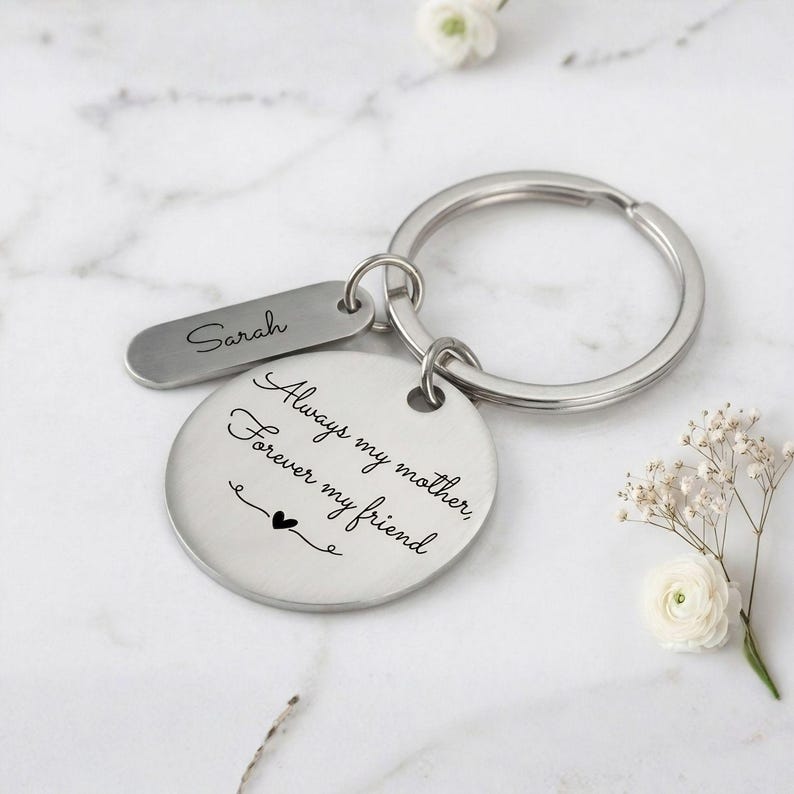 Personalized silver keychain for mom with engraved message “Always my mother, forever my friend” and custom name tag, displayed on a marble background with delicate white flowers.