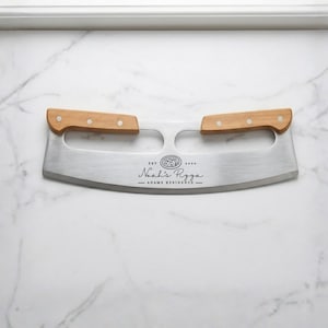 May include: A stainless steel pizza cutter with two wooden handles. The blade has the text "Noah's Pizza - Adams Residence" and a pizza graphic. The cutter is designed for slicing pizza.