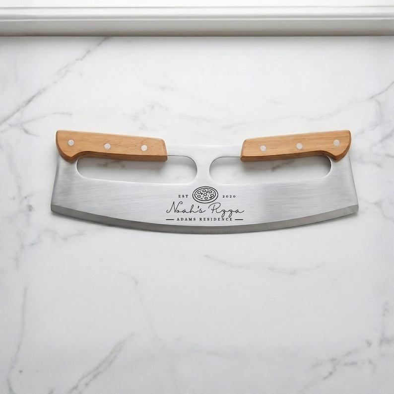 May include: A stainless steel pizza cutter with two wooden handles. The blade has the text "Noah's Pizza - Adams Residence" and a pizza graphic. The cutter is designed for slicing pizza.