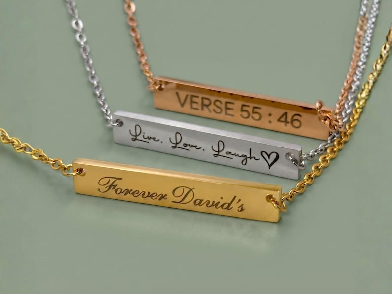 Three personalized horizontal bar necklaces in 18k Gold, Polished Silver, and Rose Gold finishes. The bars are custom engraved with a Bible verse, the phrase "Live, Love, Laugh" with a heart, and "Forever David's" in elegant script fonts.