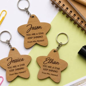 Flat lay of three personalized wooden star-shaped keychains engraved with the names "Jason," "Jessica," and "Ethan." Each keychain features the message, "YOU ARE A STAR KEEP SHINING! From Mrs. Howard." The laser-cut wooden keychains are arranged on a white and pastel green background surrounded by colorful paper clips, notebooks, and a highlighter, creating a cheerful classroom-themed display.