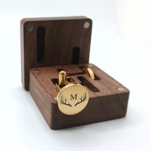 A gold-colored cufflink with the letter "M" and antler design, displayed in an open, dark brown wooden box. The box has slots for cufflinks and a ring. The cufflinks are round and shiny.