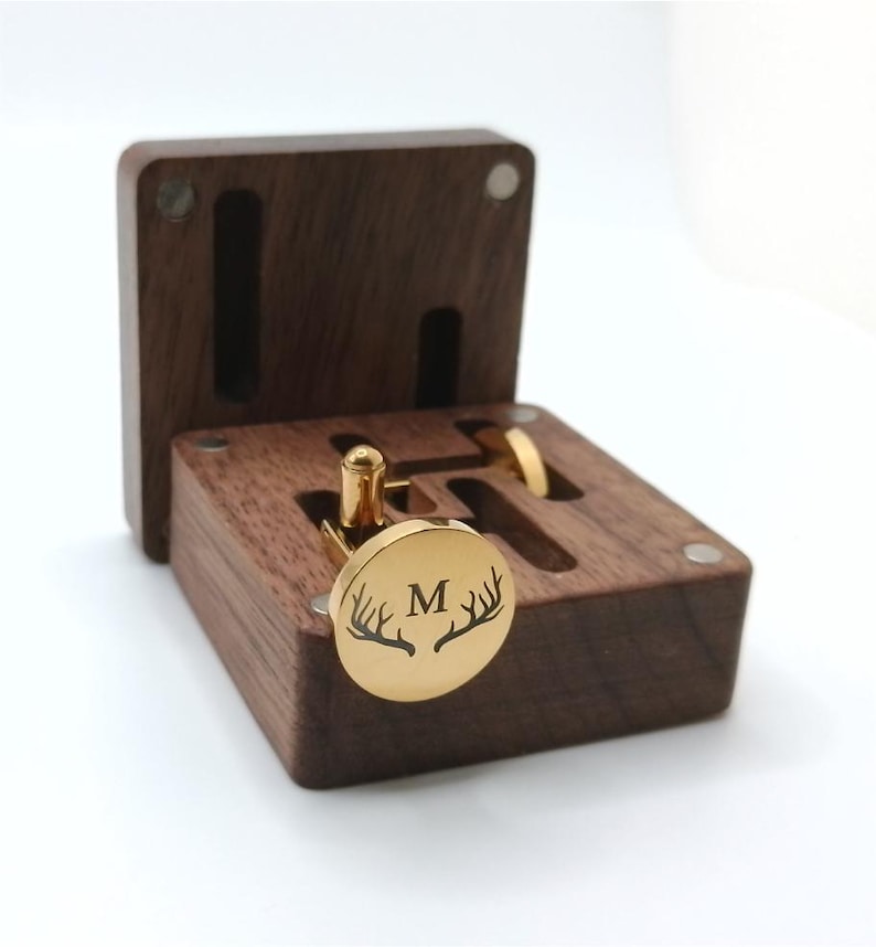 A gold-colored cufflink with the letter "M" and antler design, displayed in an open, dark brown wooden box. The box has slots for cufflinks and a ring. The cufflinks are round and shiny.