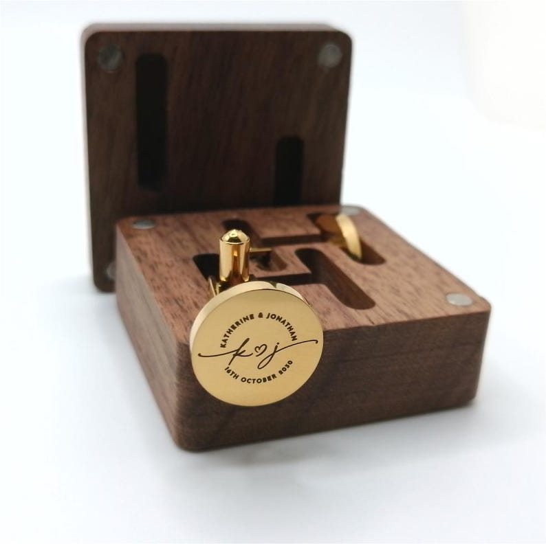 Puede incluir: Gemelos dorados con una inscripci&oacute;n personalizada, presentados en una caja de madera oscura. Los gemelos redondos est&aacute;n grabados con nombres y una fecha. La caja est&aacute; abierta, mostrando los gemelos y su almacenamiento.