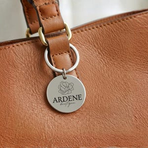 May include: A close-up of a brown leather handbag with a silver round tag attached. The tag has a floral design and the text "ARDENE boutique" in black. The bag has a brown leather strap with gold-colored hardware.