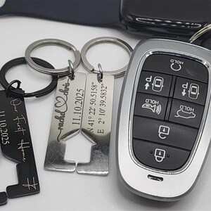 A collection of personalized keychains and a car key. The keychains include engraved text with dates and names, and a house-shaped design. The car key has a silver and black remote with multiple buttons.