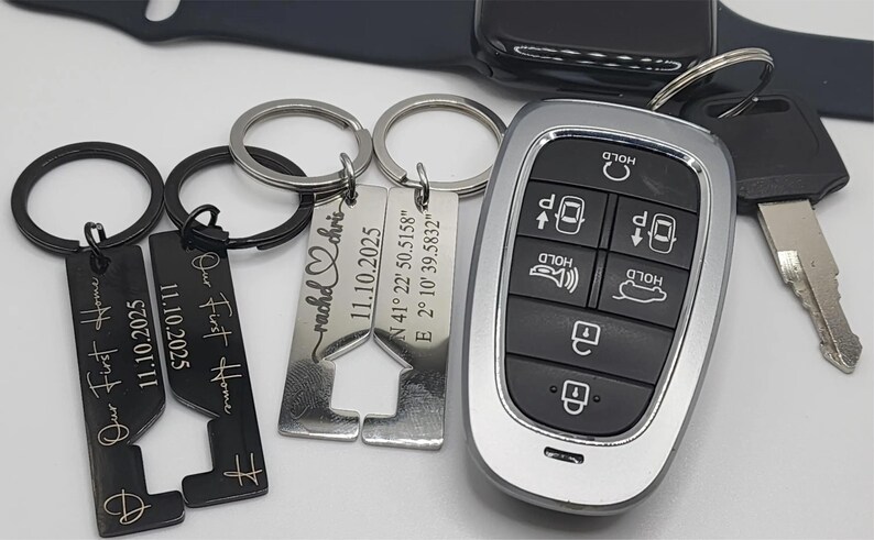A collection of personalized keychains and a car key. The keychains include engraved text with dates and names, and a house-shaped design. The car key has a silver and black remote with multiple buttons.