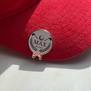 Puede incluir: Una gorra de b&eacute;isbol de color rojo brillante con un marcador de pelota de golf circular de plata sujeto a la visera. El marcador tiene una cara blanca con un gr&aacute;fico negro de dos palos de golf, una pelota y hojas de laurel, con el texto MAX BY PAR.