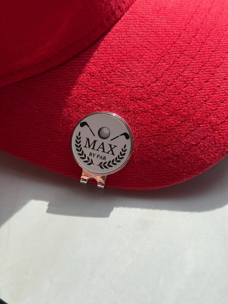 Puede incluir: Una gorra de b&eacute;isbol de color rojo brillante con un marcador de pelota de golf circular de plata sujeto a la visera. El marcador tiene una cara blanca con un gr&aacute;fico negro de dos palos de golf, una pelota y hojas de laurel, con el texto MAX BY PAR.