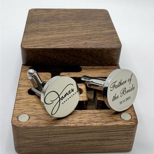 A pair of custom silver cufflinks resting in an open dark wooden box. The cufflink on the left is engraved with the name "James" in a large script font, with the word "GROOMSMAN" in a smaller block font underneath. The cufflink on the right is engraved with "Father of the Bride" in a similar script/block font combination, with the date "10.11.2025" below it.