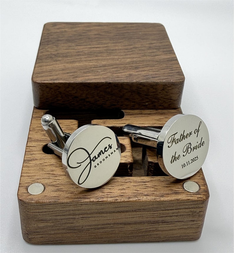 A pair of custom silver cufflinks resting in an open dark wooden box. The cufflink on the left is engraved with the name "James" in a large script font, with the word "GROOMSMAN" in a smaller block font underneath. The cufflink on the right is engraved with "Father of the Bride" in a similar script/block font combination, with the date "10.11.2025" below it.