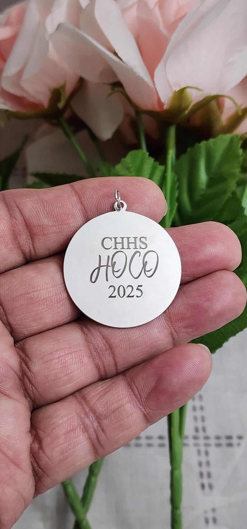 K&ouml;nnte beinhalten: Ein kreisf&ouml;rmiger silberfarbener Metallanh&auml;nger mit einer Gravur, die den Text CHHS, HOCO und 2025 vertikal untereinander zeigt. Der Anh&auml;nger verf&uuml;gt &uuml;ber einen kleinen Metallring an der Oberseite zur Befestigung und hat eine glatte Oberfl&auml;che.