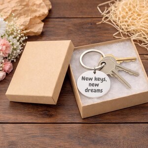 K&ouml;nnte beinhalten: Ein Schl&uuml;sselanh&auml;nger mit Schl&uuml;sseln und einem runden Anh&auml;nger in einer kleinen Geschenkbox. Auf dem Anh&auml;nger steht "New keys, new dreams". Die Box ist ge&ouml;ffnet und zeigt die Schl&uuml;ssel und den Anh&auml;nger. Ein Blumenstrau&szlig; und dekoratives Stroh befinden sich im Hintergrund.
