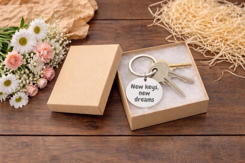 K&ouml;nnte beinhalten: Ein Schl&uuml;sselanh&auml;nger mit Schl&uuml;sseln und einem runden Anh&auml;nger in einer kleinen Geschenkbox. Auf dem Anh&auml;nger steht "New keys, new dreams". Die Box ist ge&ouml;ffnet und zeigt die Schl&uuml;ssel und den Anh&auml;nger. Ein Blumenstrau&szlig; und dekoratives Stroh befinden sich im Hintergrund.
