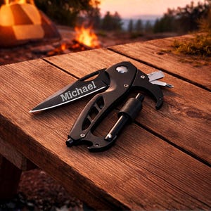 A black multi-tool knife with a carabiner is shown lying flat on a wooden surface. The blade is partially open and features the word "Michael" engraved on it. The handle is ergonomic and has several functions, including a Phillips-head screwdriver, a flathead screwdriver, and a hexagonal wrench. The multi-tool is positioned diagonally, with the carabiner on the right and the knife blade on the left. The lighting is bright and even.