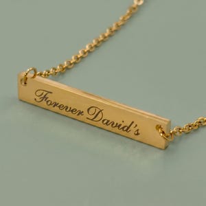 A gold-colored necklace with a rectangular pendant. The pendant is engraved with the words "Forever David's" in a cursive font. The necklace chain is delicate and has a beaded design.