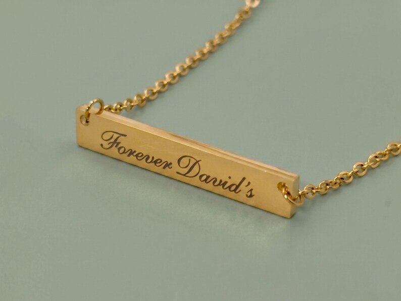 A gold-colored necklace with a rectangular pendant. The pendant is engraved with the words "Forever David's" in a cursive font. The necklace chain is delicate and has a beaded design.