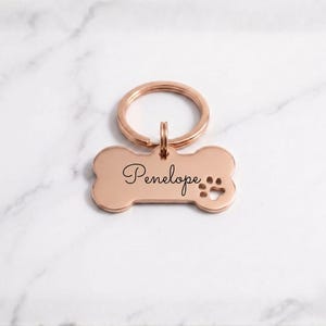 Personalized rose gold dog tag with engraved name Penelope in cursive. Custom bone-shaped brass pet ID tag with paw print cutout and gold keyring on a marble surface.