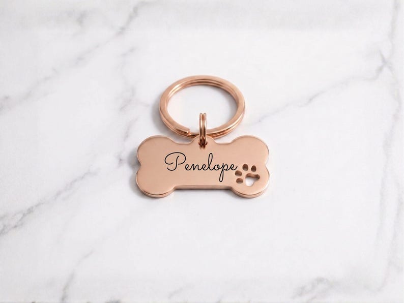 Personalized rose gold dog tag with engraved name Penelope in cursive. Custom bone-shaped brass pet ID tag with paw print cutout and gold keyring on a marble surface.