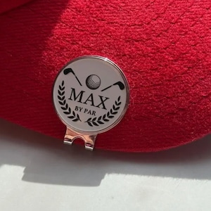 Personalized magnetic golf ball marker with hat clip featuring crossed golf clubs, a golf ball, laurel wreath, and the name "MAX BY PAR," attached to the brim of a red golf cap. Silver metal marker with white enamel finish, ideal as a custom golfer gift and golf accessory.