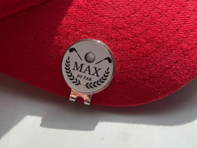 Personalized magnetic golf ball marker with hat clip featuring crossed golf clubs, a golf ball, laurel wreath, and the name "MAX BY PAR," attached to the brim of a red golf cap. Silver metal marker with white enamel finish, ideal as a custom golfer gift and golf accessory.