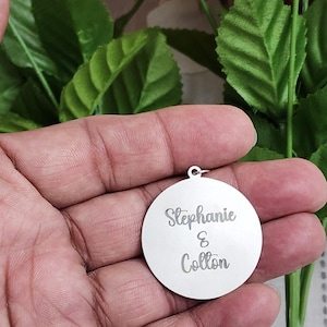 K&ouml;nnte beinhalten: Ein runder Anh&auml;nger aus silberfarbenem Metall mit der eingravierten Aufschrift Stephanie & Colton in Schreibschrift. Der Anh&auml;nger verf&uuml;gt &uuml;ber einen kleinen Verbindungsring an der Oberseite zur Befestigung und hat eine glatte, reflektierende Oberfl&auml;che.