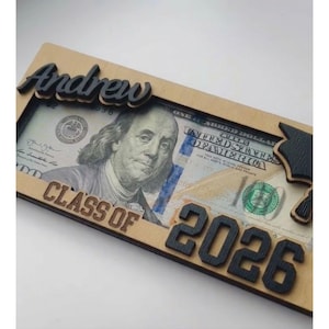Close-up angled view of a personalized laser-engraved wooden graduation money holder reading "Andrew – Class of 2026," with a $100 bill tucked inside the open slot, showing the raised black script name, bold engraved year numbers, laser-cut graduation cap detail on the right corner, and natural birch wood grain texture against a clean white background.